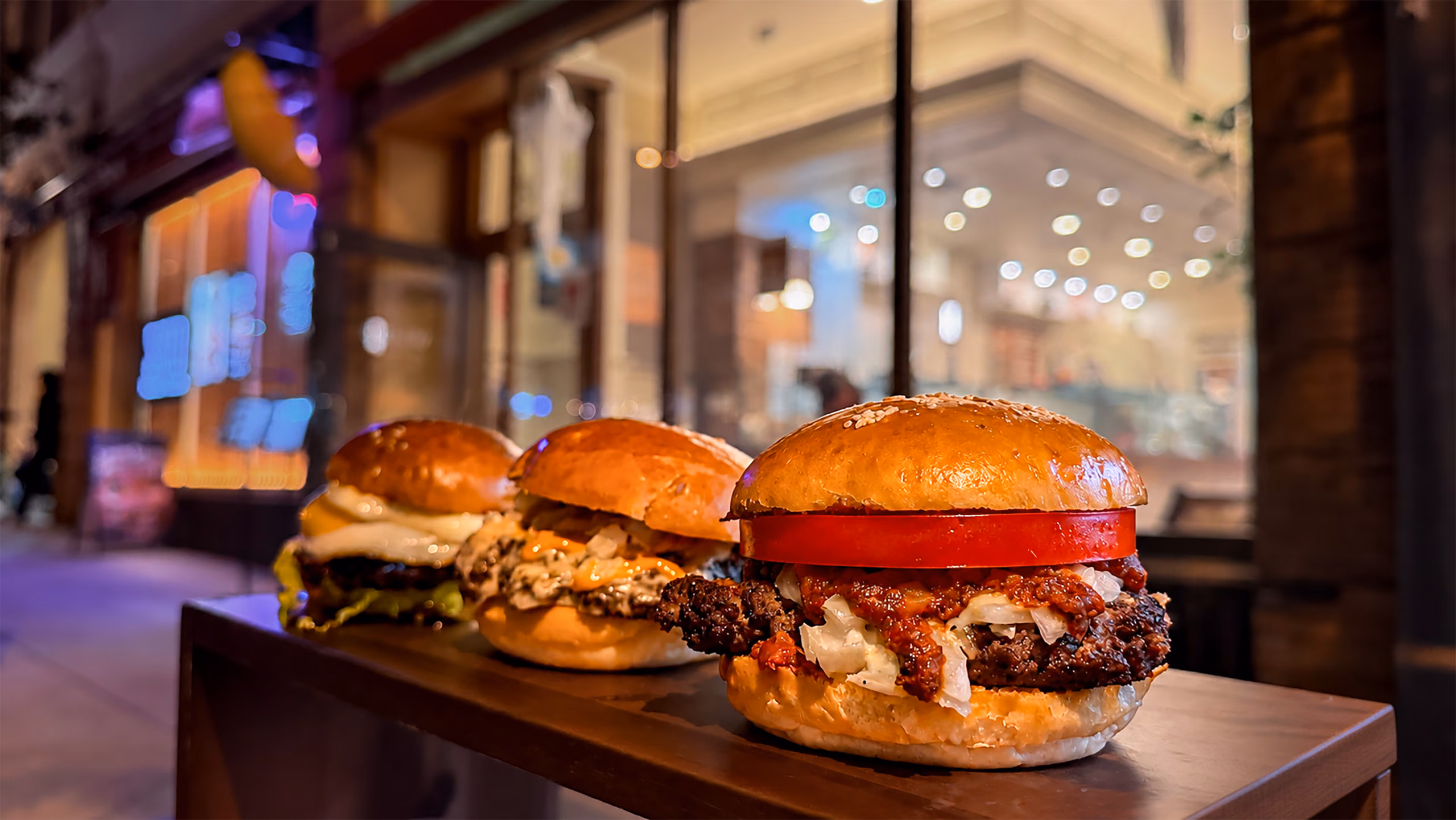 Three burgers on sesame brioche buns lined up on a wooden ledge outside the café at night, the nearest topped with a thick tomato slice and relish, the lit storefront blurred behind them.