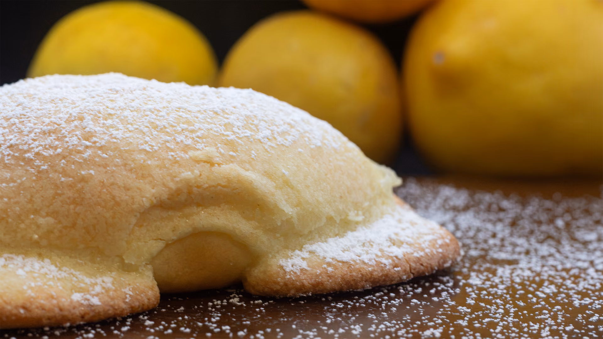A sugar-dusted bun with pale lemon curd easing from its side, whole lemons blurred behind it.